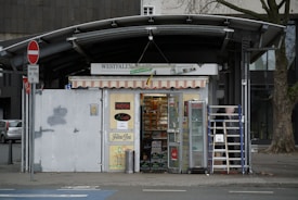 A small kiosk located on a street corner with a variety of goods displayed inside. The kiosk has a colorful striped awning and signage on the walls. A ladder is positioned against the storefront, and various advertisements are visible. The exterior includes a street sign and a tree nearby.