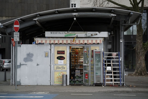 A small kiosk located on a street corner with a variety of goods displayed inside. The kiosk has a colorful striped awning and signage on the walls. A ladder is positioned against the storefront, and various advertisements are visible. The exterior includes a street sign and a tree nearby.