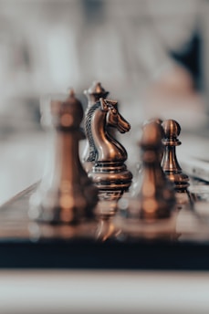 A close-up view of several chess pieces, including a knight, positioned on a chessboard. The scene is focused and detailed, with the pieces exhibiting a metallic sheen against a blurred background.