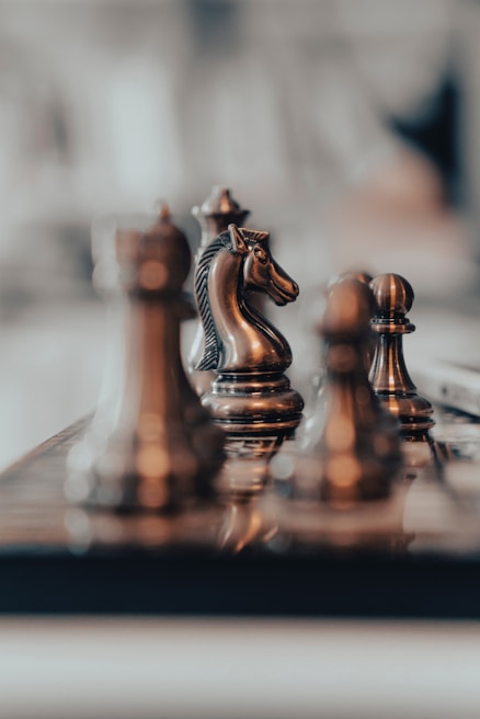 A close-up view of several chess pieces, including a knight, positioned on a chessboard. The scene is focused and detailed, with the pieces exhibiting a metallic sheen against a blurred background.