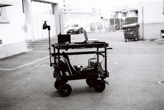 A black and white image depicting a multi-level trolley with various electronic equipment, cables, and a mounted camera in an empty parking area. In the background, a couple of cars and a dumpster can be seen near buildings.