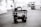 A black and white image depicting a multi-level trolley with various electronic equipment, cables, and a mounted camera in an empty parking area. In the background, a couple of cars and a dumpster can be seen near buildings.