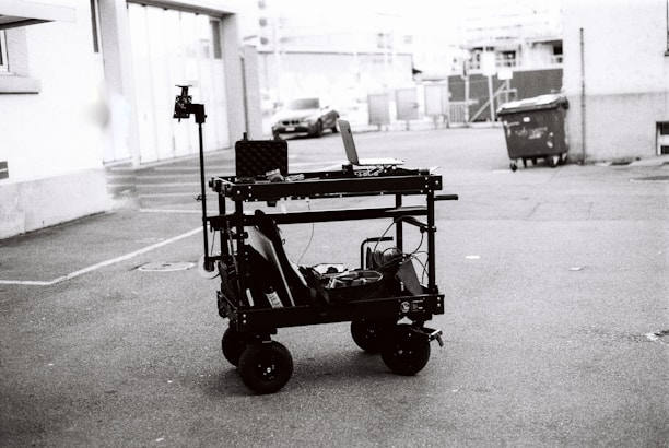 A black and white image depicting a multi-level trolley with various electronic equipment, cables, and a mounted camera in an empty parking area. In the background, a couple of cars and a dumpster can be seen near buildings.