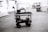 A black and white image depicting a multi-level trolley with various electronic equipment, cables, and a mounted camera in an empty parking area. In the background, a couple of cars and a dumpster can be seen near buildings.