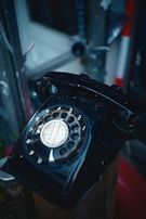 A vintage black rotary dial telephone is shown, positioned on a wooden surface. The dial numbers are clearly visible and the surroundings are slightly blurred, indicating an indoor setting with various metallic and wooden elements.