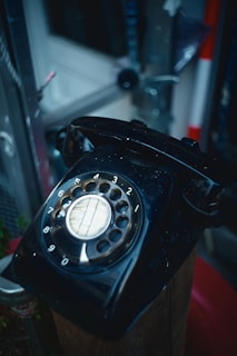 A vintage black rotary dial telephone is shown, positioned on a wooden surface. The dial numbers are clearly visible and the surroundings are slightly blurred, indicating an indoor setting with various metallic and wooden elements.