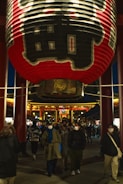 A large, traditional red lantern with black and gold details hangs prominently in a temple setting, surrounded by bustling people. The structure is illuminated, indicating an evening or night scene. Visitors, many wearing face masks, walk beneath the lantern and around the temple, suggesting a lively but serene atmosphere.
