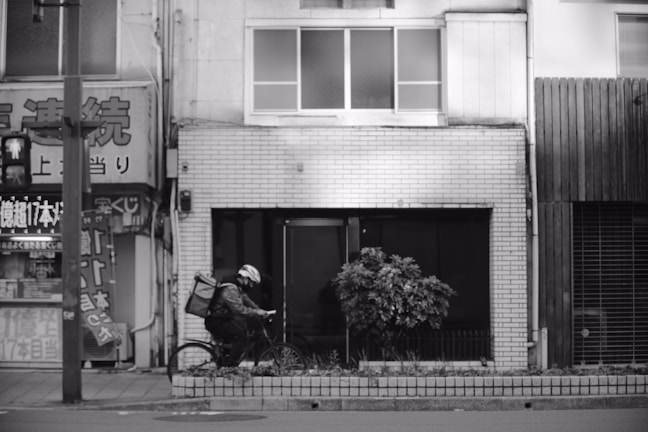 A cyclist wearing a helmet rides a bicycle along a city street. They carry a large backpack, possibly for deliveries. The background features a row of urban storefronts, with signs and advertisements written in Japanese. The photograph is in black and white, emphasizing the textures of the urban environment and the rider.