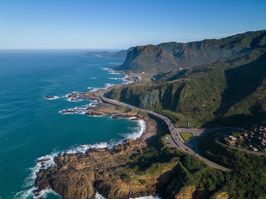 A scenic coastal landscape featuring a winding road along rugged cliffs overlooking the ocean. The shoreline is dotted with rocky formations and waves gently lapping against them. There are green hills rising above the road, with scattered buildings in the distance. The sky is clear and blue, adding to the serene atmosphere.
