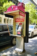A public payphone booth with a pink canopy displaying a Telstra logo and offering free Wi-Fi is situated on a city street. Nearby, parked cars and trees are visible, suggesting an urban environment. The booth features a metallic payphone unit with a keypad and display.