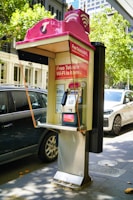 A public payphone booth with a pink canopy displaying a Telstra logo and offering free Wi-Fi is situated on a city street. Nearby, parked cars and trees are visible, suggesting an urban environment. The booth features a metallic payphone unit with a keypad and display.