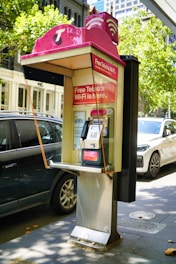 A public payphone booth with a pink canopy displaying a Telstra logo and offering free Wi-Fi is situated on a city street. Nearby, parked cars and trees are visible, suggesting an urban environment. The booth features a metallic payphone unit with a keypad and display.