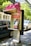 A public payphone booth with a pink canopy displaying a Telstra logo and offering free Wi-Fi is situated on a city street. Nearby, parked cars and trees are visible, suggesting an urban environment. The booth features a metallic payphone unit with a keypad and display.