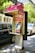 A public payphone booth with a pink canopy displaying a Telstra logo and offering free Wi-Fi is situated on a city street. Nearby, parked cars and trees are visible, suggesting an urban environment. The booth features a metallic payphone unit with a keypad and display.
