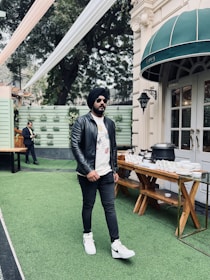 A man wearing a black leather jacket, white graphic t-shirt, black pants, and white sneakers walks on a green artificial grass surface. He is also wearing sunglasses and a black turban. Nearby, there is a buffet table with dishes and cutlery set up outside a building with a green awning.