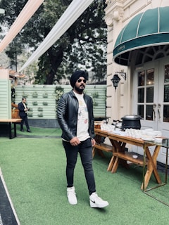 A man wearing a black leather jacket, white graphic t-shirt, black pants, and white sneakers walks on a green artificial grass surface. He is also wearing sunglasses and a black turban. Nearby, there is a buffet table with dishes and cutlery set up outside a building with a green awning.