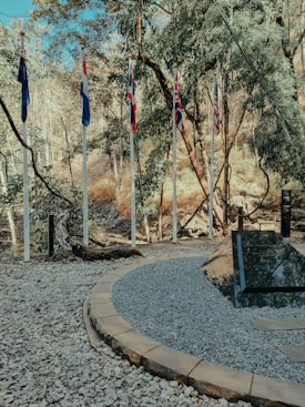 A row of flagpoles displaying several national flags stands in a natural, wooded setting. The flags include designs with red, white, and blue colors. The area is surrounded by trees, and the ground is covered in gravel with a circular path. A large rock with an inscription is present near the flags.