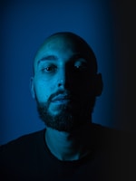A portrait of a bearded man with a shaved head is lit dramatically from one side, resulting in a strong contrast between light and shadow. The background is a solid, deep blue, enhancing the somber tone of the photograph.
