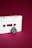 A white audio cassette tape is displayed against a deep magenta background. A black and textured object that resembles the shape of a guitar pick is placed below one of the cassette's empty spools, giving it a somewhat anthropomorphic appearance.