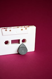 A white audio cassette tape is displayed against a deep magenta background. A black and textured object that resembles the shape of a guitar pick is placed below one of the cassette's empty spools, giving it a somewhat anthropomorphic appearance.