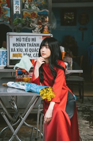 A person in a red outfit sits at a metal table in an outdoor setting, holding a bouquet of yellow flowers. Behind them is a sign with Vietnamese writing, a colorful decorative panel, and small figurines.