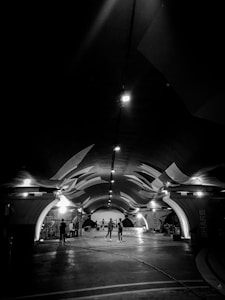 A black and white image of an arched underground space with a sleek, modern design. Illuminated by a series of overhead lights, the space has uniquely shaped ceilings with angular patterns. Several people are present, some standing and others seemingly engaged in various activities. The words 'LEARN' and 'SHARE' can be seen on the walls.