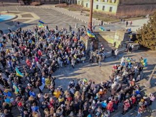 A large group of people gathered in a public square, holding flags and banners with blue and yellow colors. The crowd appears to be engaged in a protest or rally, with a strong presence of Ukrainian flags. The scene is set in an urban environment with steps and a historic building nearby.