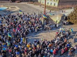 A large group of people gathered in a public square, holding flags and banners with blue and yellow colors. The crowd appears to be engaged in a protest or rally, with a strong presence of Ukrainian flags. The scene is set in an urban environment with steps and a historic building nearby.