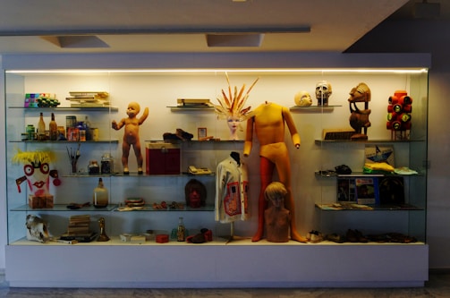 A display case filled with a variety of eclectic objects, including a mannequin, dolls, books, bottles, hats, masks, and figurines. The arrangement is colorful and visually interesting with a mix of modern and vintage items.
