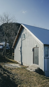 A small, corrugated metal house stands in a grassy area, surrounded by bare trees. The structure has a gabled roof, a few windows, and a door with some shading on the concrete foundation. The sky is clear, and the image has a cool, shadowy tone reflecting the winter season.