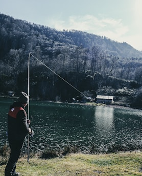 A person wearing a beanie and jacket is fishing by a calm lake surrounded by dense forest. The water reflects the sky, and there is a small cabin or structure visible near the water's edge. Tall trees on surrounding hills provide a scenic backdrop.