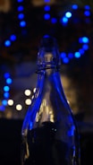 A glass bottle is silhouetted against a backdrop of blurry blue and white bokeh lights, creating a contrast between the sharp outline of the bottle and the soft, diffused lights in the background.