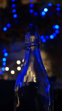 A glass bottle is silhouetted against a backdrop of blurry blue and white bokeh lights, creating a contrast between the sharp outline of the bottle and the soft, diffused lights in the background.