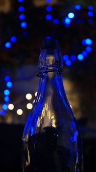 A glass bottle is silhouetted against a backdrop of blurry blue and white bokeh lights, creating a contrast between the sharp outline of the bottle and the soft, diffused lights in the background.