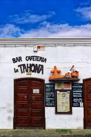 Front facade of a bar and cafe named 'La Tahona' with a white wall, dark brown wooden doors, and a decorative sign above the entrance. The sign includes large clay jugs and a small guitar placed above a shelf. Below the decorations, there are menus and various signs displayed on the wall. The sky is bright blue with some clouds.