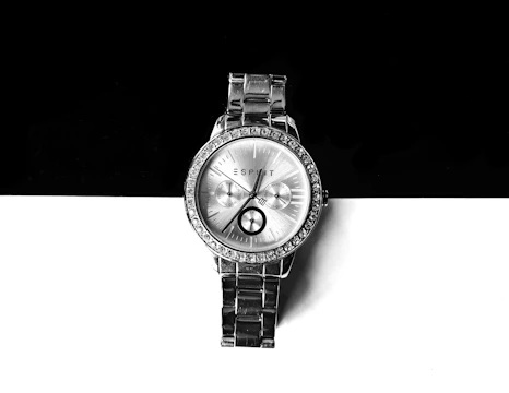 A silver wristwatch with a metallic bracelet is positioned against a contrasting background of black and white. The watch face is adorned with small crystals around the bezel and features three sub-dials. Roman numerals and simple batons mark the hour, and the brand name is printed at the top.