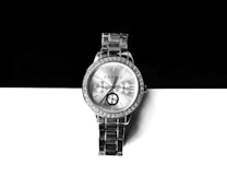 A silver wristwatch with a metallic bracelet is positioned against a contrasting background of black and white. The watch face is adorned with small crystals around the bezel and features three sub-dials. Roman numerals and simple batons mark the hour, and the brand name is printed at the top.