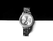 A silver wristwatch with a metallic bracelet is positioned against a contrasting background of black and white. The watch face is adorned with small crystals around the bezel and features three sub-dials. Roman numerals and simple batons mark the hour, and the brand name is printed at the top.