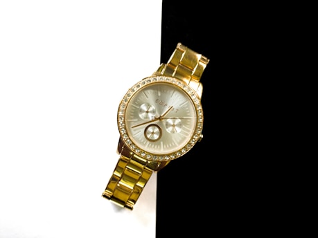 A gold wristwatch with a metallic band and a round face featuring a crystal-studded bezel is positioned against a stark black and white background. The watch face shows three sub-dials and has a sophisticated, luxurious appearance with gold detailing.