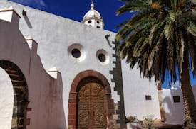 A whitewashed building with a dome and Romanesque architectural features, including arched windows and a large, ornate wooden door. A tall palm tree stands nearby, casting its shadow against the wall. The sky is clear and blue, adding a vibrant contrast to the building’s earthy tones.