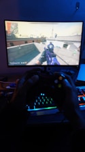 A person is engaged in playing a first-person shooter game on a computer monitor. The screen shows a game screen with a map and a player holding a weapon. Below the screen, a pair of hands holds a black gaming controller illuminated by colorful RGB lights from a keyboard.