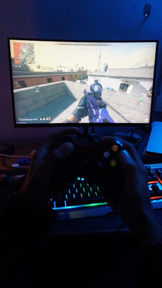 A person is engaged in playing a first-person shooter game on a computer monitor. The screen shows a game screen with a map and a player holding a weapon. Below the screen, a pair of hands holds a black gaming controller illuminated by colorful RGB lights from a keyboard.