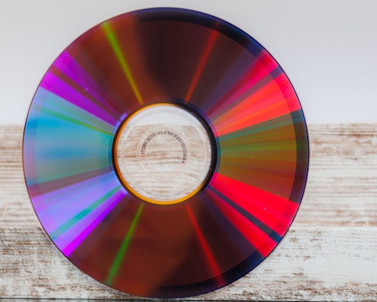A close-up of a compact disc resting on a wooden surface, reflecting a spectrum of colors including red, blue, green, and purple. The disc's reflective surface creates a vivid and dynamic appearance, with the concentric patterns emphasizing the CD's design.