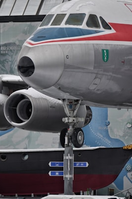 The image features the front section of an aircraft with a focus on the cockpit and the nose. The aircraft is predominantly silver with red and blue stripes. Snow is visible on parts of the aircraft. The landing gear is extended and clearly visible, positioned over directional signs at the base. A backdrop with geographical names and elevation markers is present, hinting at a museum or themed environment.