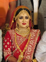 A bride is adorned in traditional red attire with intricate gold embroidery. She wears elaborate gold jewelry, including a necklace, earrings, and a headpiece. Her makeup is bold, featuring red lipstick and dark eye cosmetics.