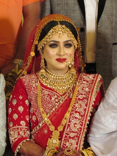 A bride is adorned in traditional red attire with intricate gold embroidery. She wears elaborate gold jewelry, including a necklace, earrings, and a headpiece. Her makeup is bold, featuring red lipstick and dark eye cosmetics.