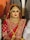A bride is adorned in traditional red attire with intricate gold embroidery. She wears elaborate gold jewelry, including a necklace, earrings, and a headpiece. Her makeup is bold, featuring red lipstick and dark eye cosmetics.
