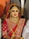 A bride is adorned in traditional red attire with intricate gold embroidery. She wears elaborate gold jewelry, including a necklace, earrings, and a headpiece. Her makeup is bold, featuring red lipstick and dark eye cosmetics.