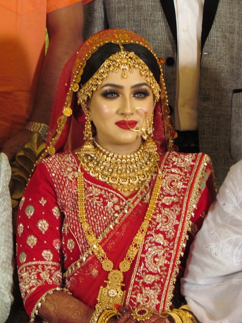 A bride is adorned in traditional red attire with intricate gold embroidery. She wears elaborate gold jewelry, including a necklace, earrings, and a headpiece. Her makeup is bold, featuring red lipstick and dark eye cosmetics.