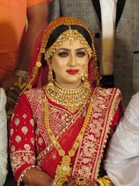 A bride is adorned in traditional red attire with intricate gold embroidery. She wears elaborate gold jewelry, including a necklace, earrings, and a headpiece. Her makeup is bold, featuring red lipstick and dark eye cosmetics.